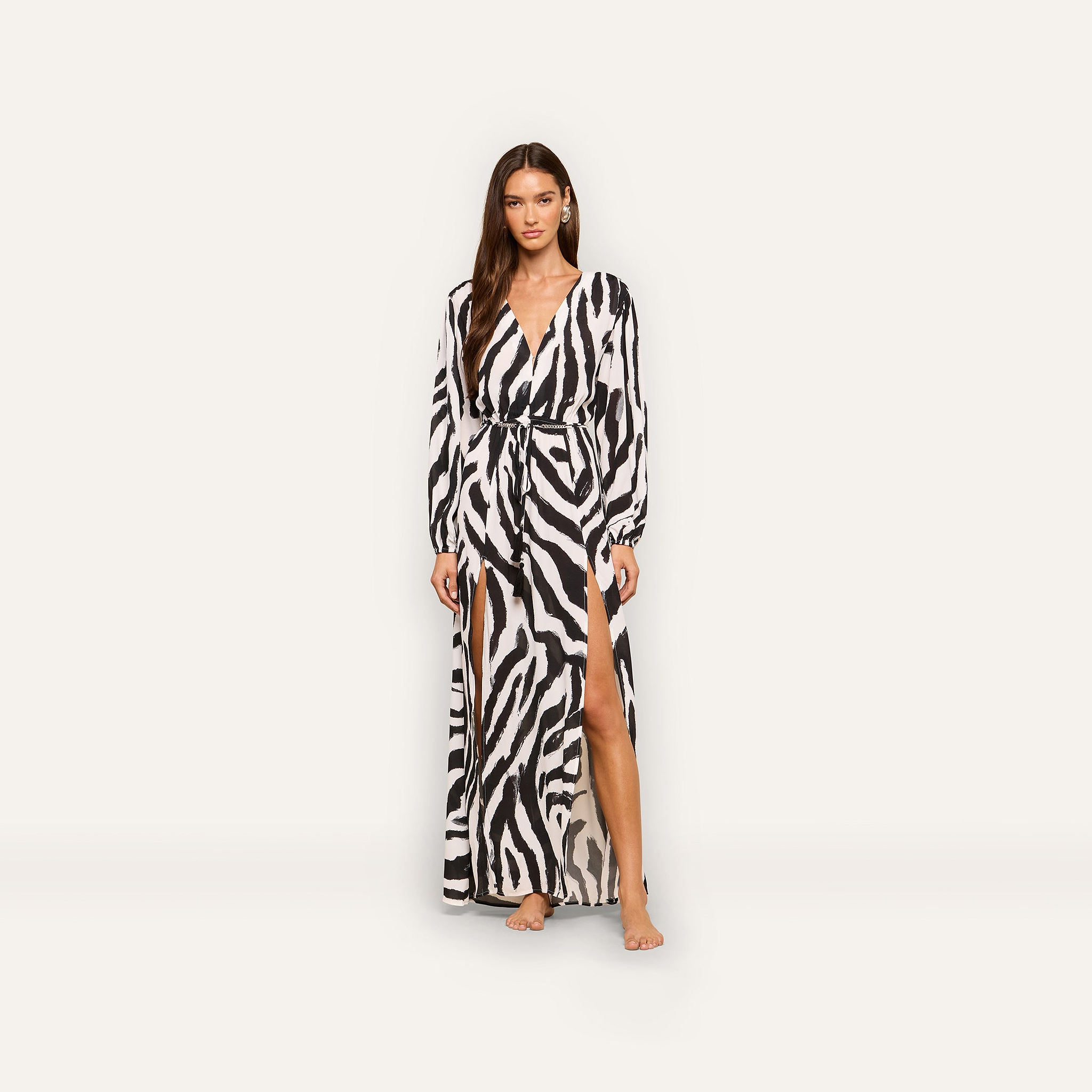 A woman stands barefoot in the Sara Maxi Cover-Up Dress, which features a zebra print, long sleeves, a deep V-neck, a belted waist, and high front slits, set against a plain white background.