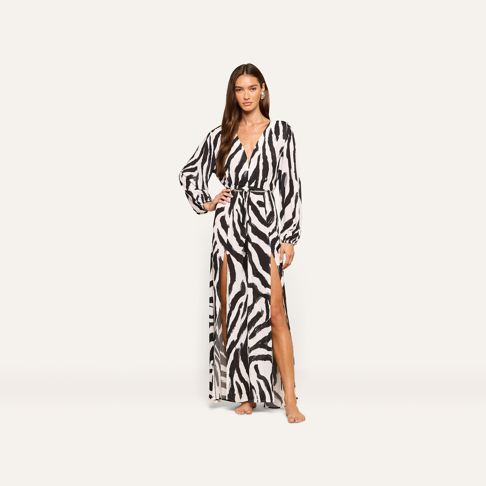 A woman wearing the Sara Maxi Cover-Up Dress, featuring a black-and-white zebra print with long sleeves and a high slit, stands barefoot against a plain off-white background.