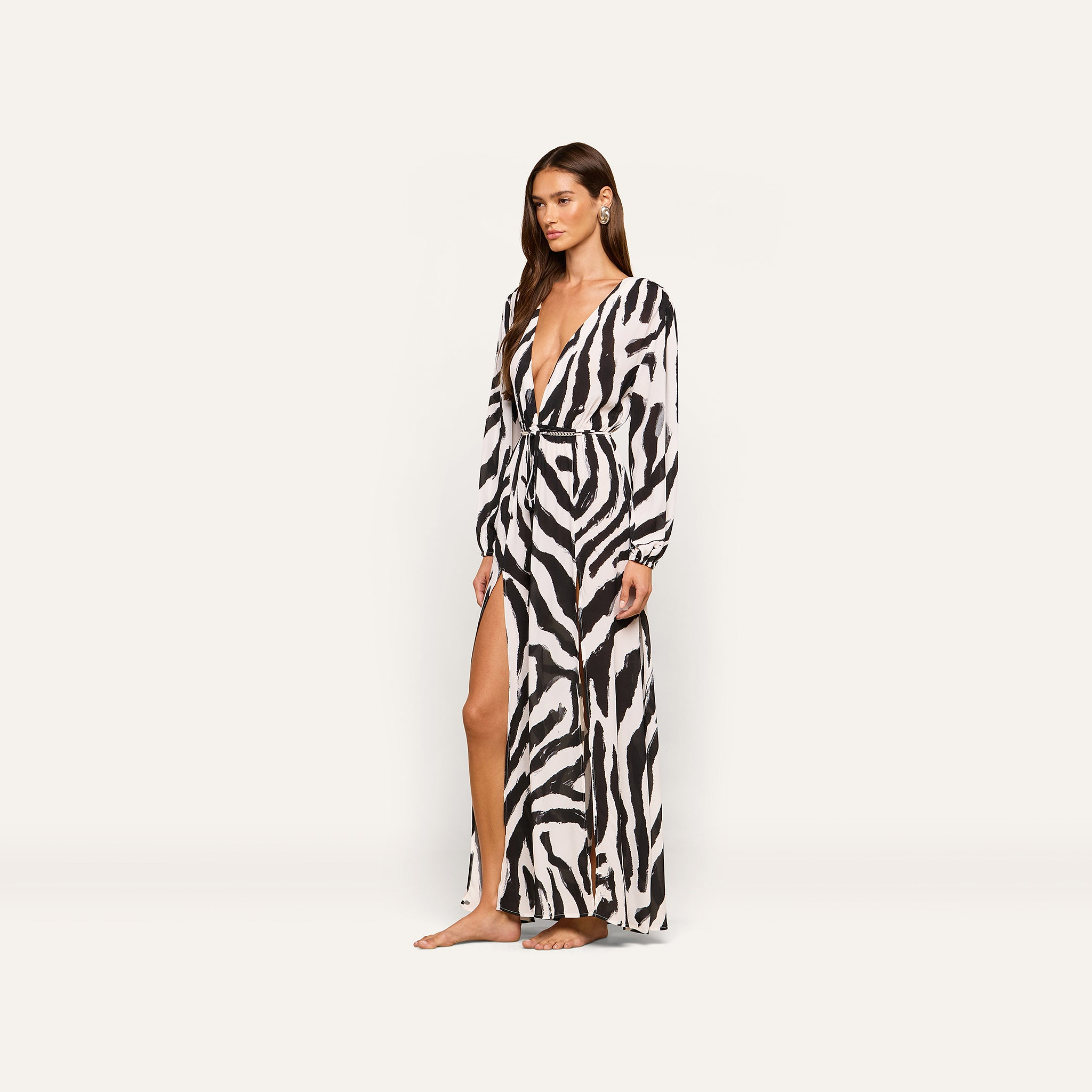 A woman models the Sara Maxi Cover-Up Dress, featuring a long zebra print design with a deep V-neck, long sleeves, and a high slit, as she stands barefoot against a plain light background.