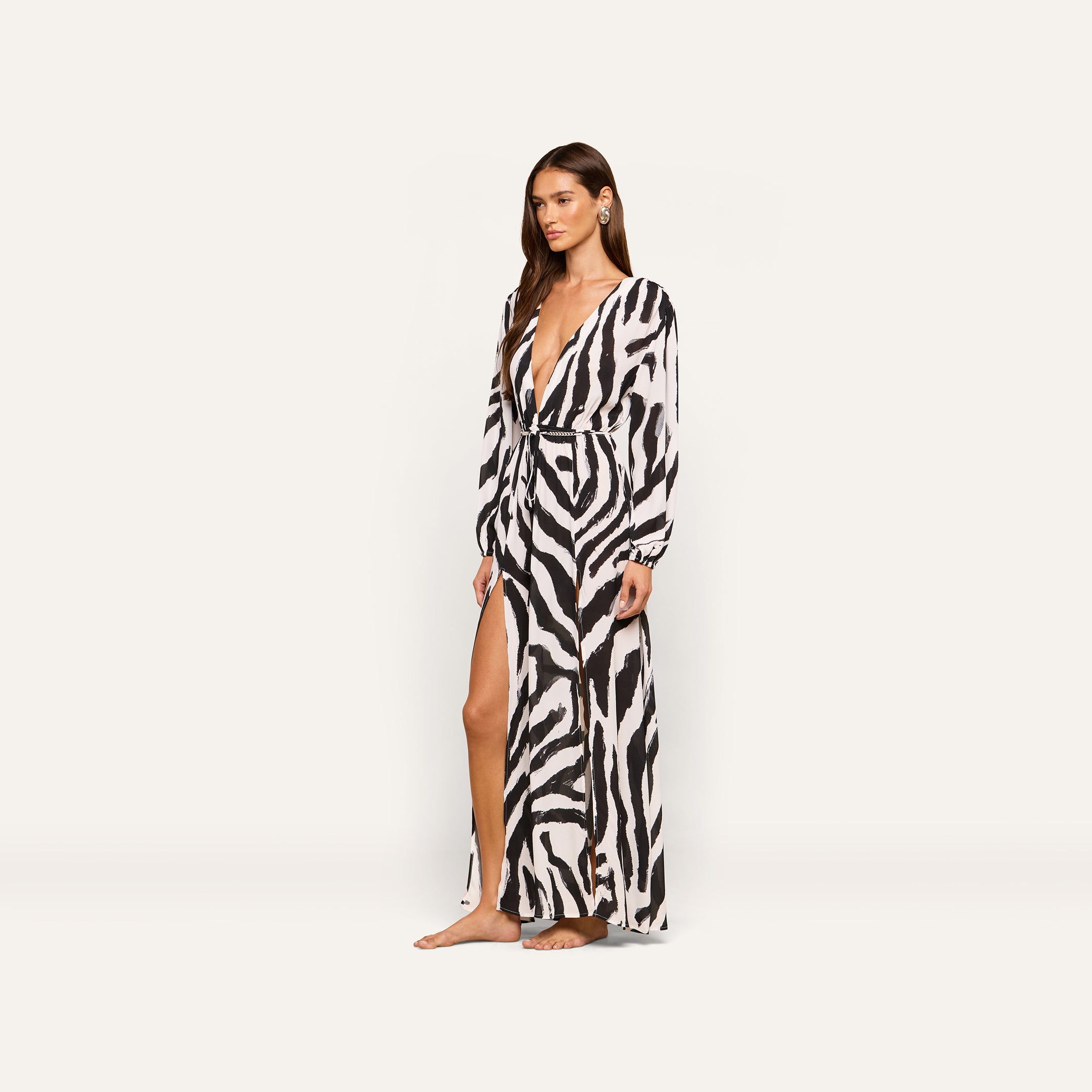 A woman models the Sara Maxi Cover-Up Dress, featuring a long zebra print design with a deep V-neck, long sleeves, and a high slit, as she stands barefoot against a plain light background.