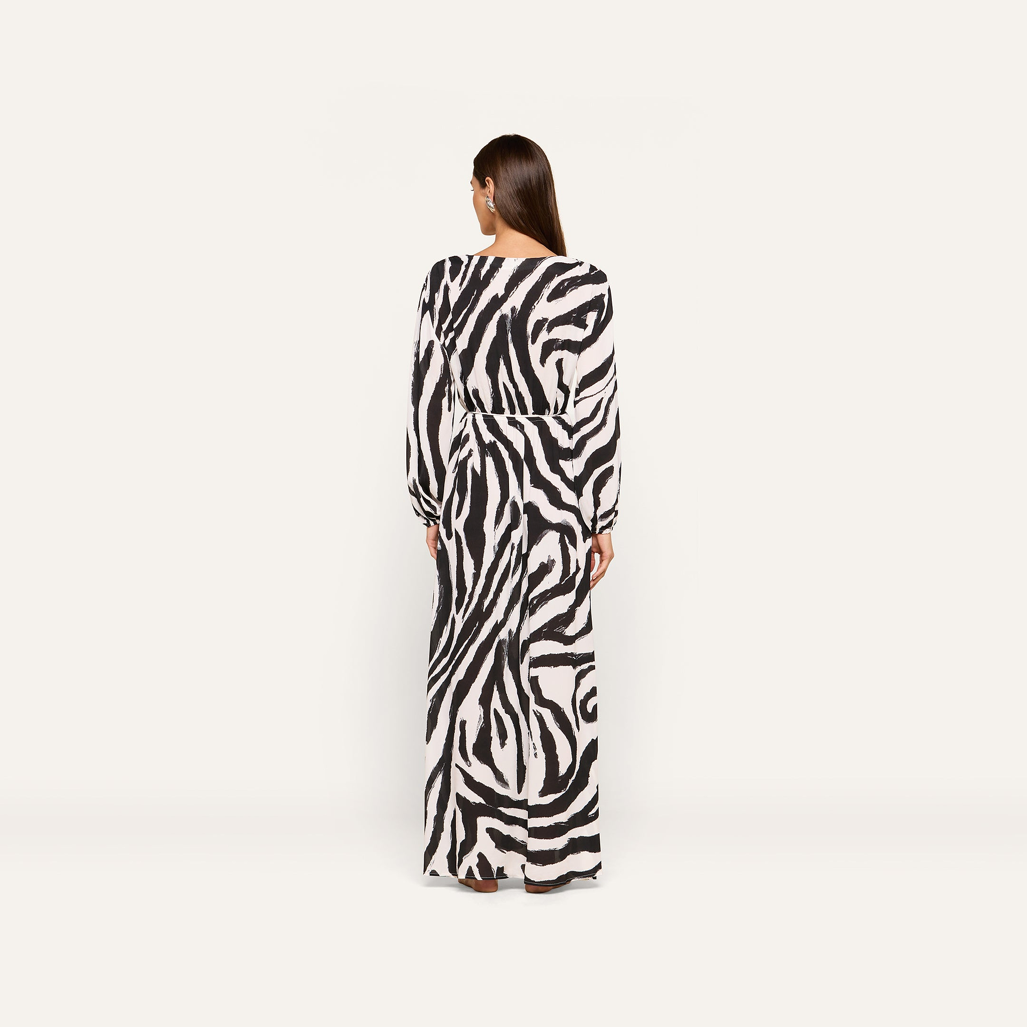 A person stands with their back to the camera, wearing the long-sleeved Sara Maxi Cover-Up Dress in a zebra print. The background is plain and light-colored.