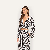 Woman wearing the Ambala Cover-Up Top, a black and white zebra-print piece with an island vibes design, featuring a deep V-neck and long sleeves. She pairs it with matching pants and poses with one hand in her pocket against a plain background.