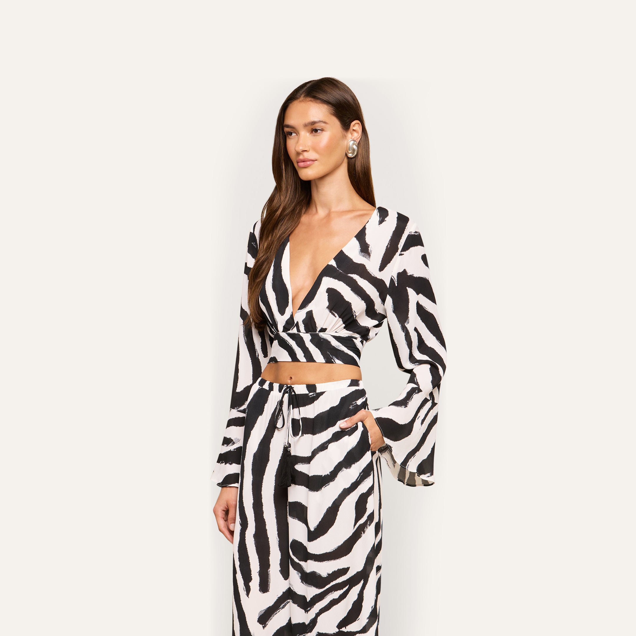Woman wearing the Ambala Cover-Up Top, a black and white zebra-print piece with an island vibes design, featuring a deep V-neck and long sleeves. She pairs it with matching pants and poses with one hand in her pocket against a plain background.