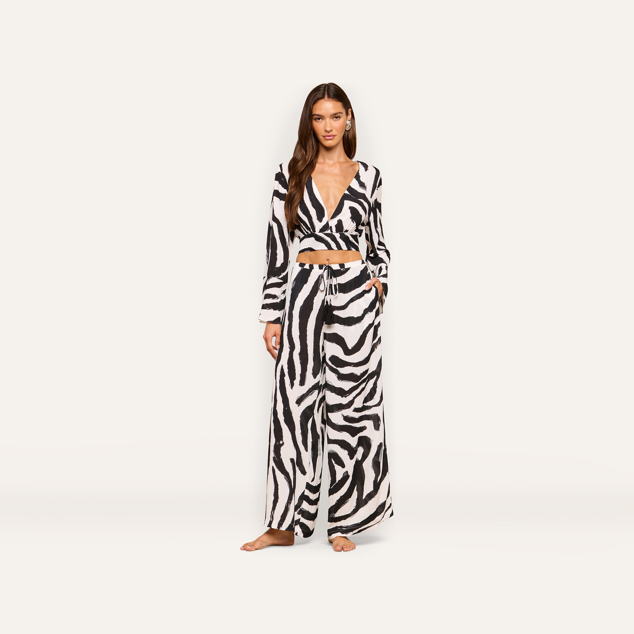 A woman stands facing forward wearing the Ambala Cover-Up Top, a matching black and white zebra print long-sleeve crop top paired with wide-leg pants. She has long brown hair and is barefoot against a plain, light background.