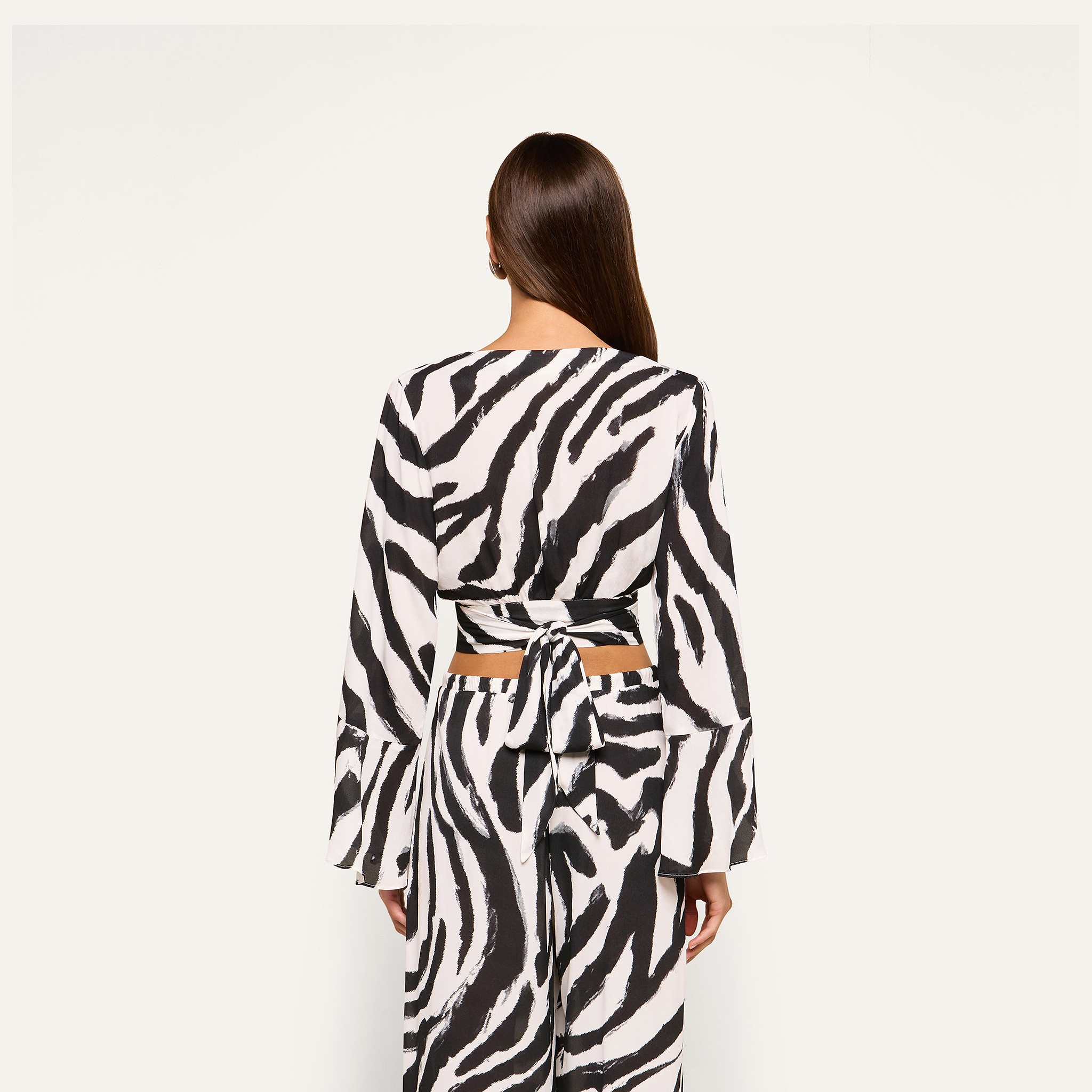 A woman with long brown hair stands facing away, wearing the Ambala Cover-Up Top—a black and white zebra print piece featuring long sleeves and a tie-wrap closure at the waist—paired with matching pants. The background is plain white.