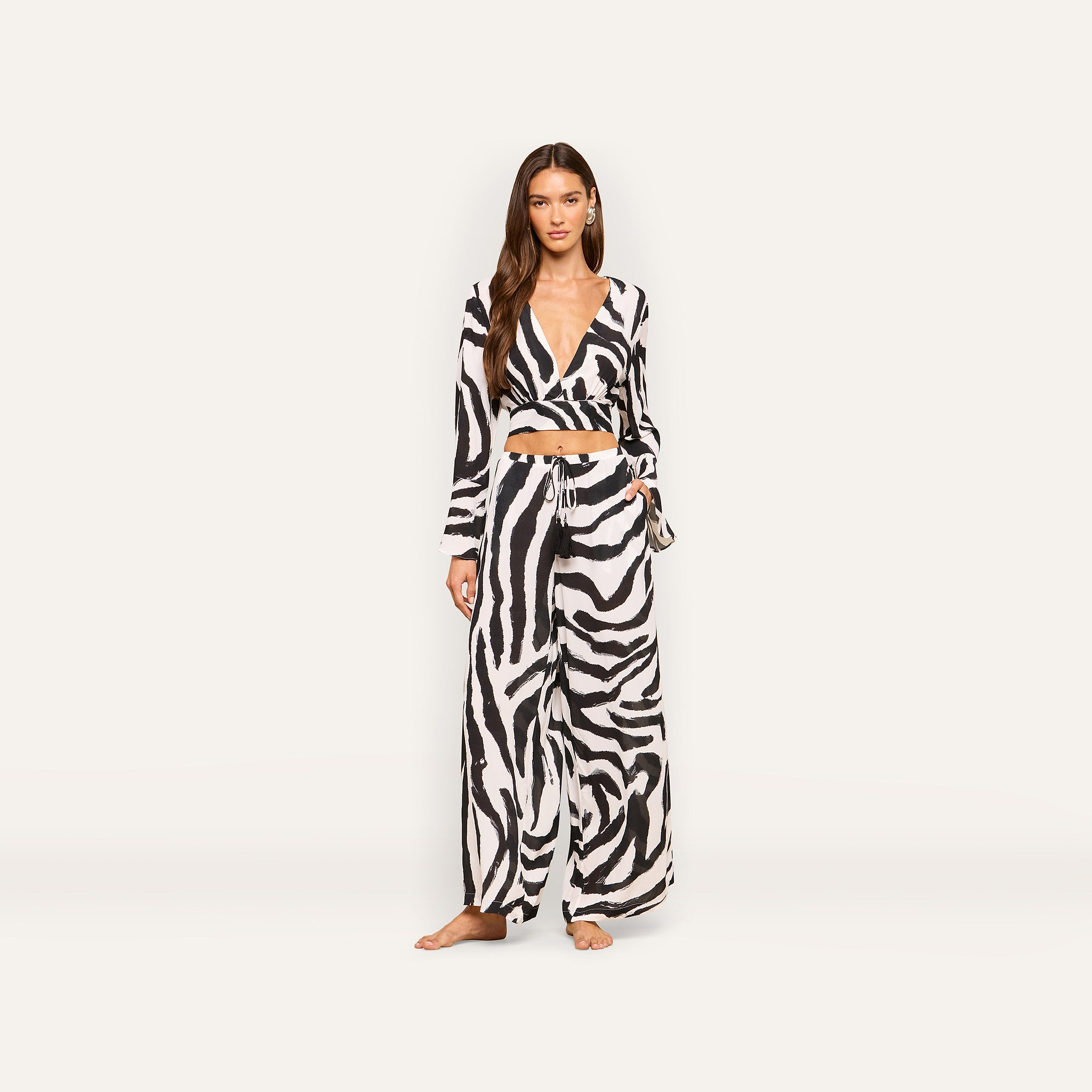 A woman stands facing forward against a plain light background, wearing the Geraldine Cover-Up Pant in a zebra-print matching set with a long-sleeve crop top and wide-leg resort wear pants.