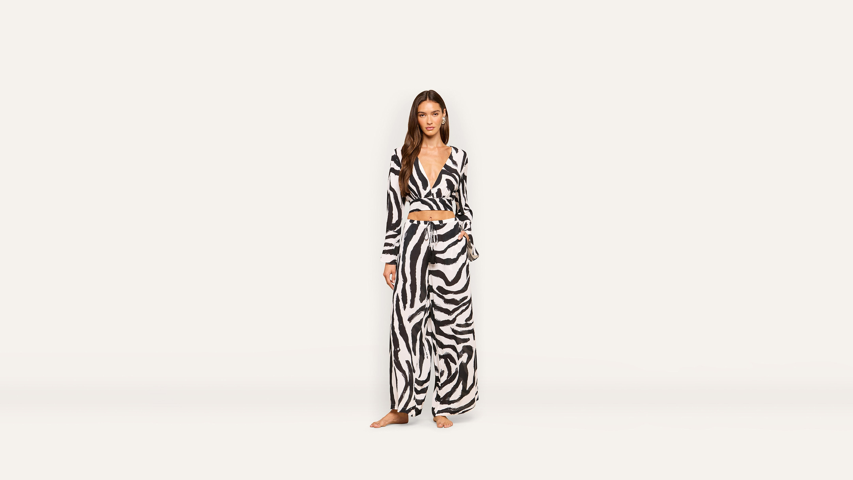 A woman stands barefoot wearing a chic black and white zebra print long-sleeve crop top and wide-leg pants against a plain light background.