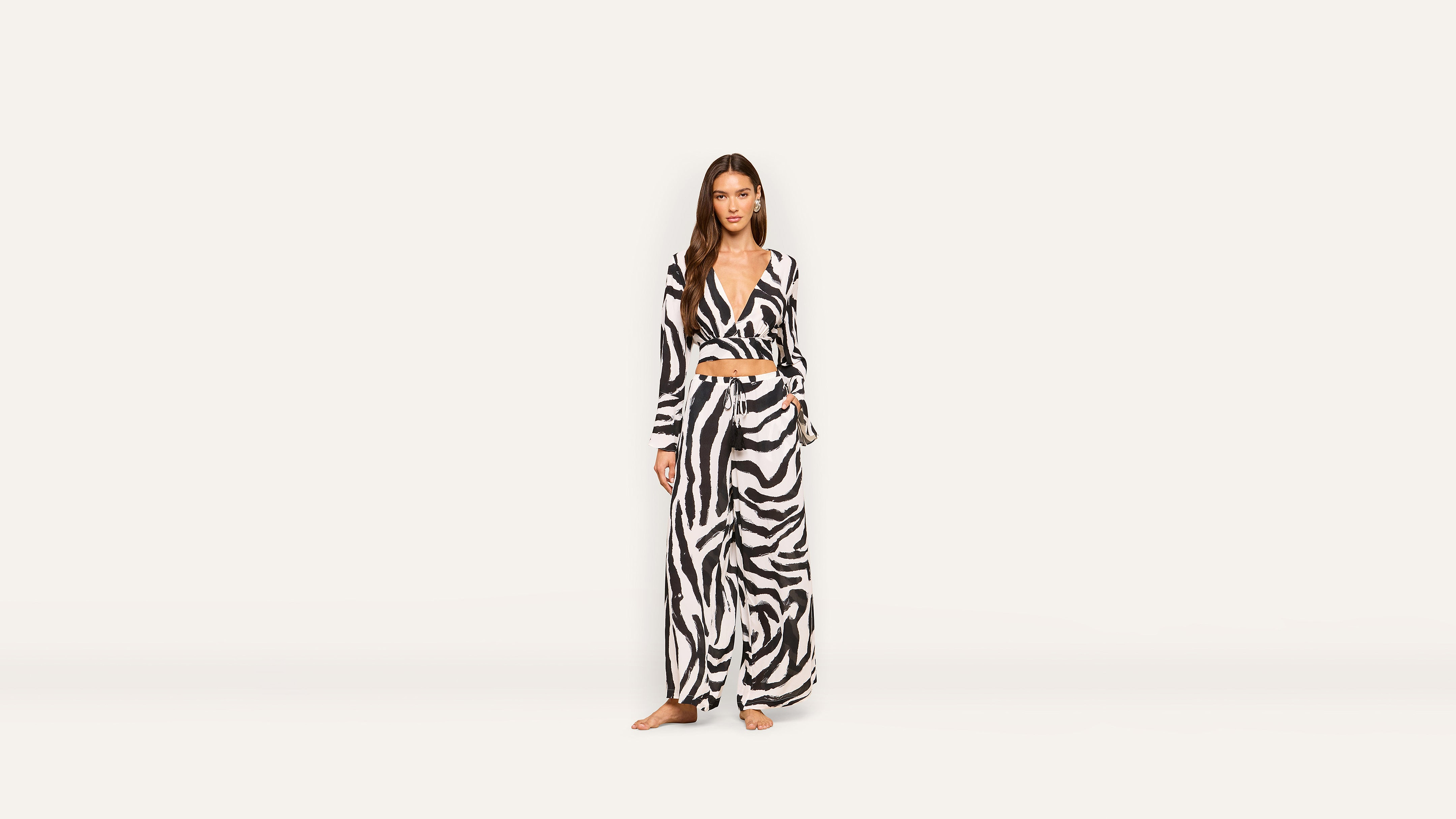 A woman stands barefoot wearing a chic black and white zebra print long-sleeve crop top and wide-leg pants against a plain light background.
