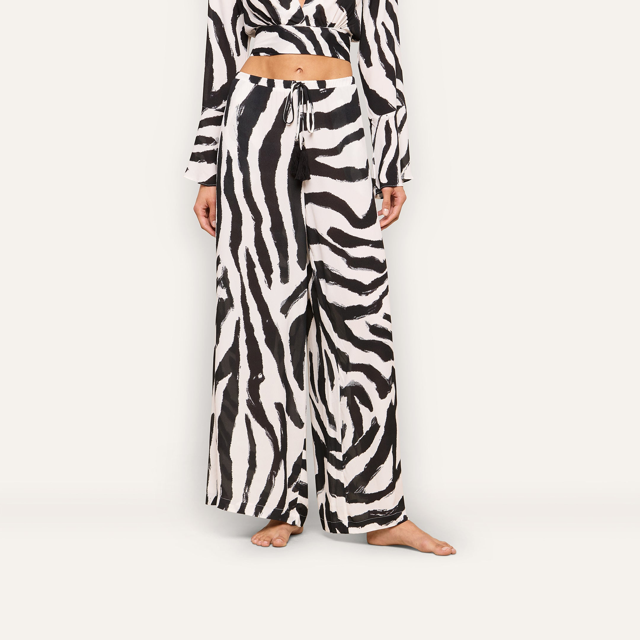 A woman models the Geraldine Cover-Up Pant featuring a zebra print, paired with a matching cropped top. She stands barefoot against a plain background, with her head cropped out of the frame.