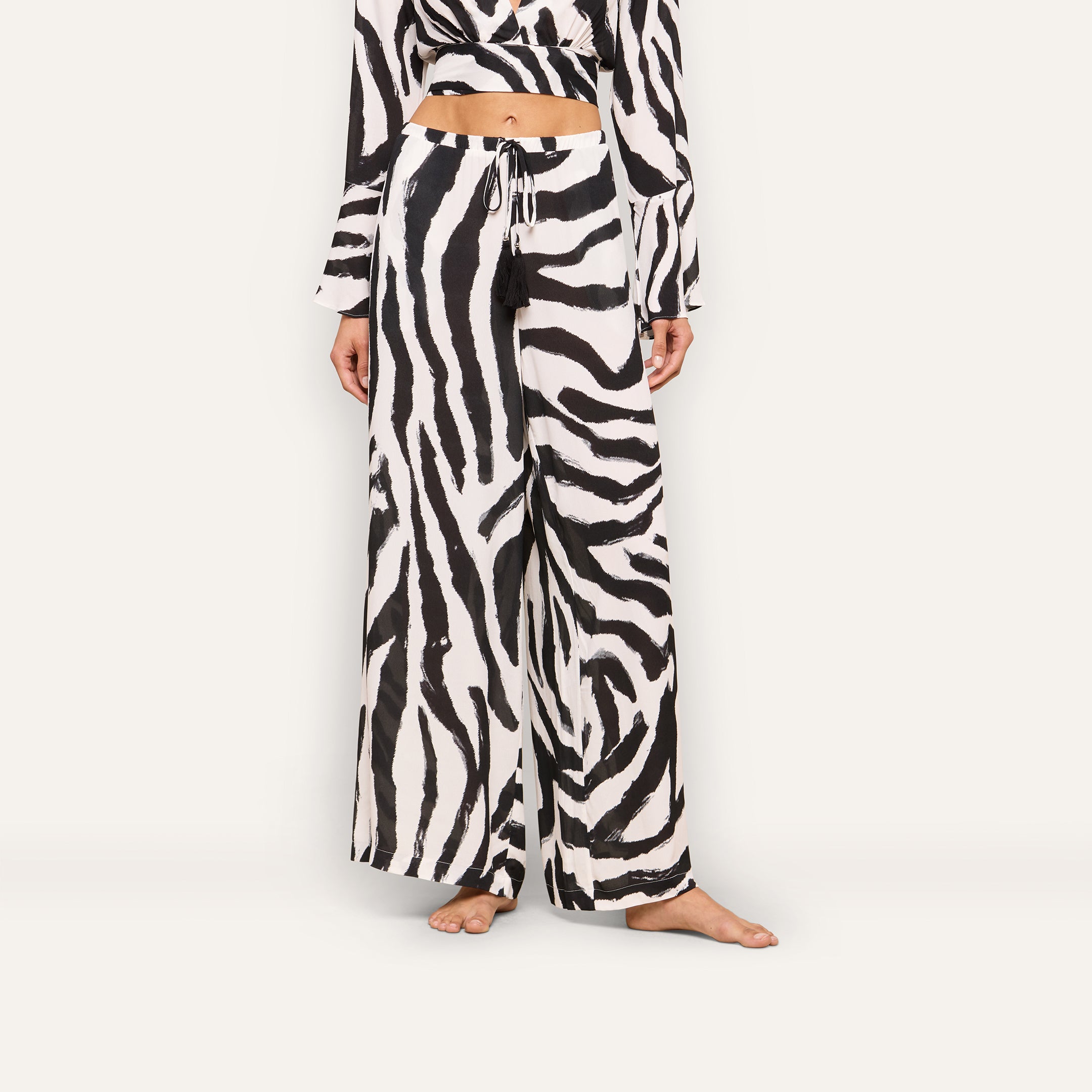 A woman models the Geraldine Cover-Up Pant featuring a zebra print, paired with a matching cropped top. She stands barefoot against a plain background, with her head cropped out of the frame.