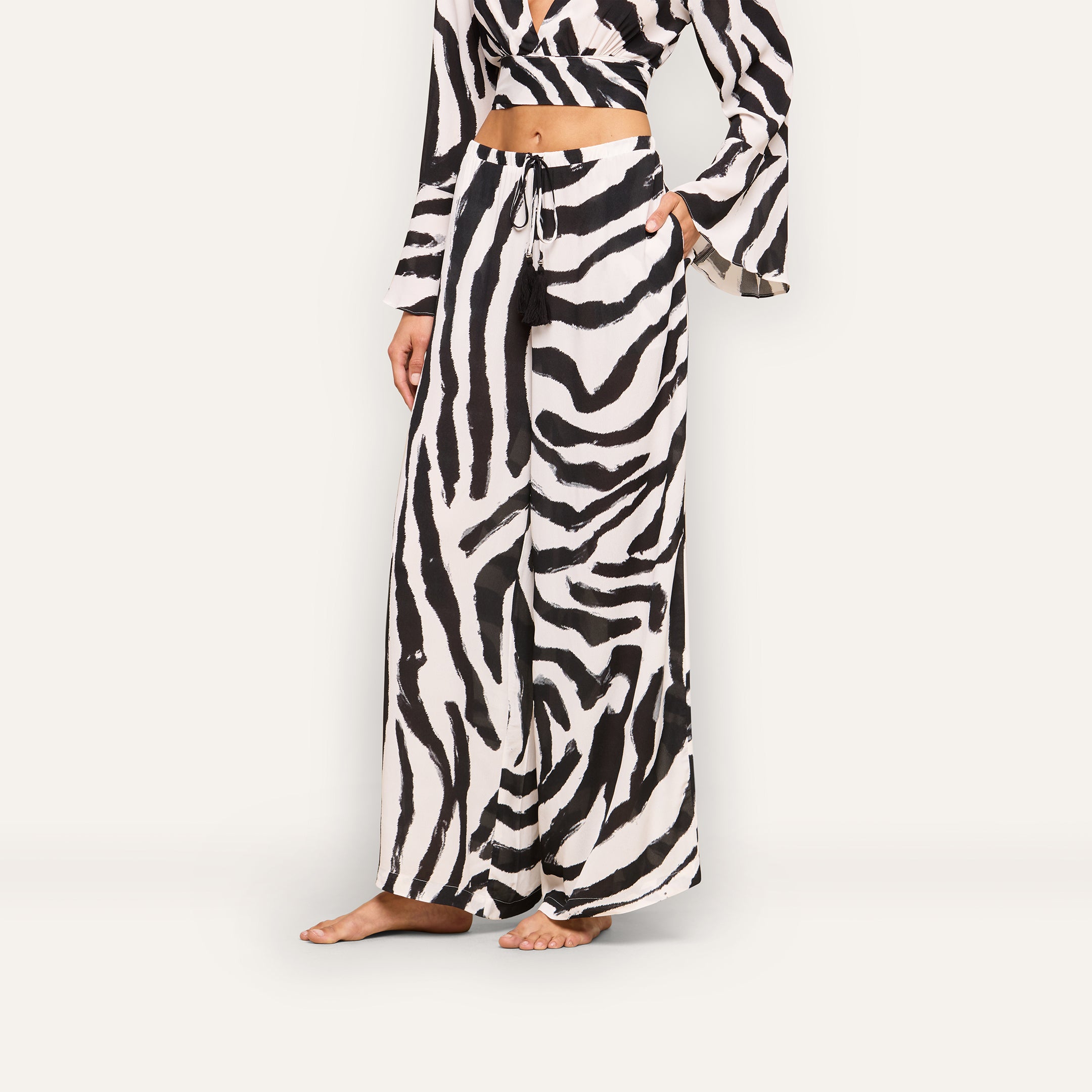 A person stands barefoot against a plain background wearing the Geraldine Cover-Up Pant in a zebra print, paired with a matching long-sleeve crop top. The person’s face is not visible.