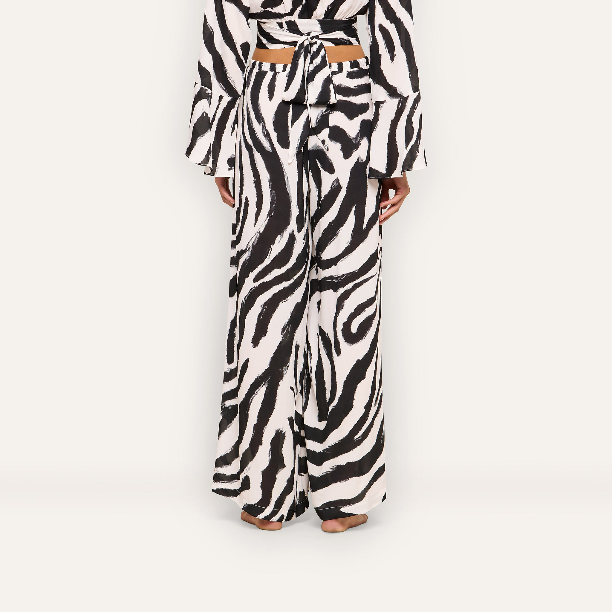 A person stands barefoot, seen from behind, wearing the Geraldine Cover-Up Pant—wide-leg zebra print pants—paired with a matching long-sleeve top that ties at the back.