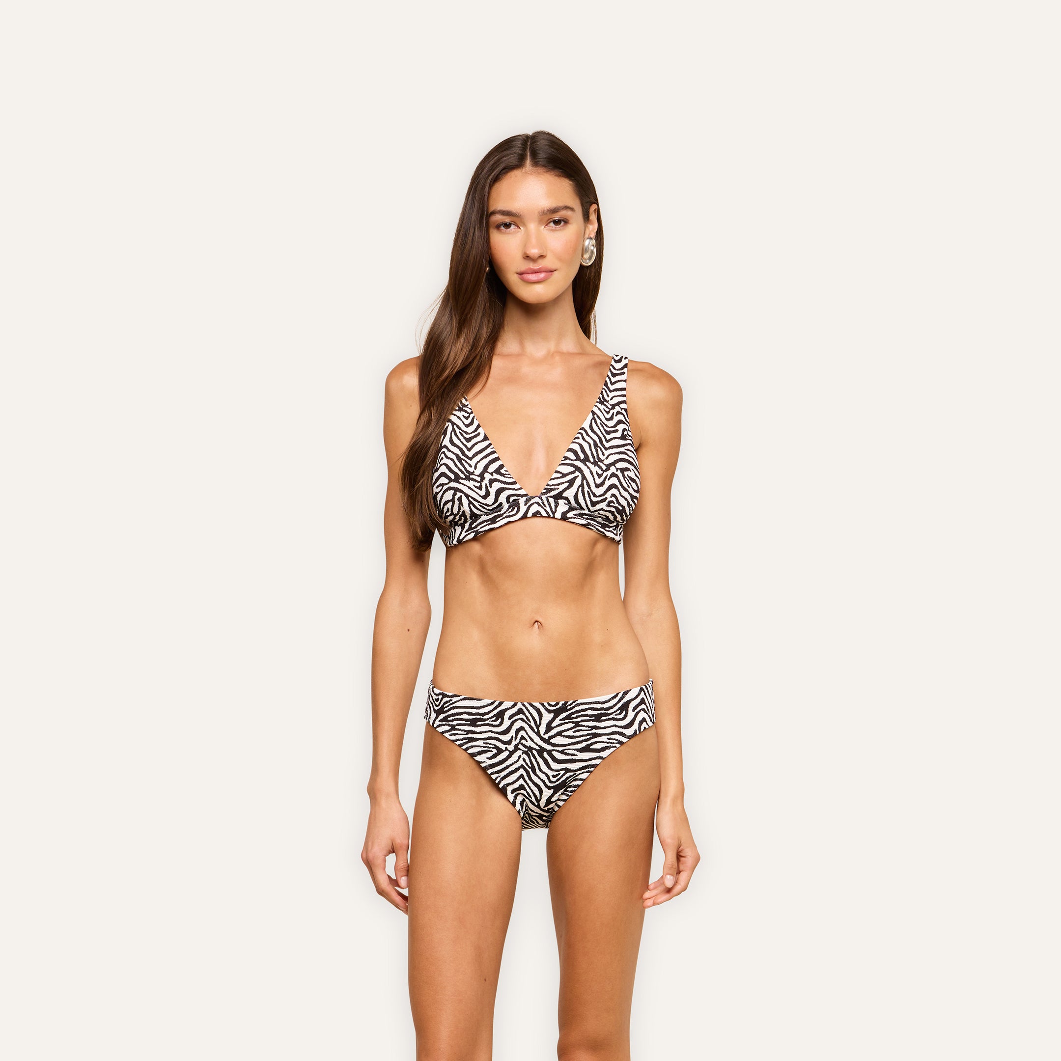 Woman stands facing forward in a zebra print Lexie Bikini Top with matching bottoms against a plain light background. She has long, wavy brown hair and a neutral expression.
