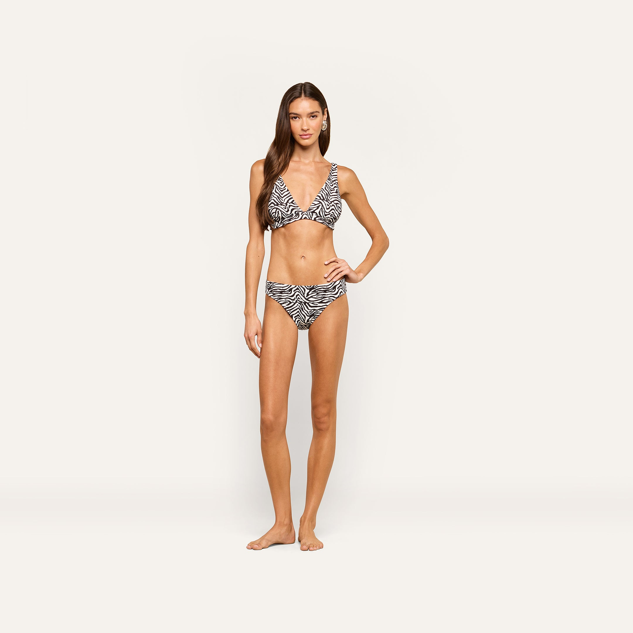 A woman with long brown hair stands barefoot and facing forward against a plain light background, wearing the Lexie Bikini Top in a zebra print, with a neutral expression.