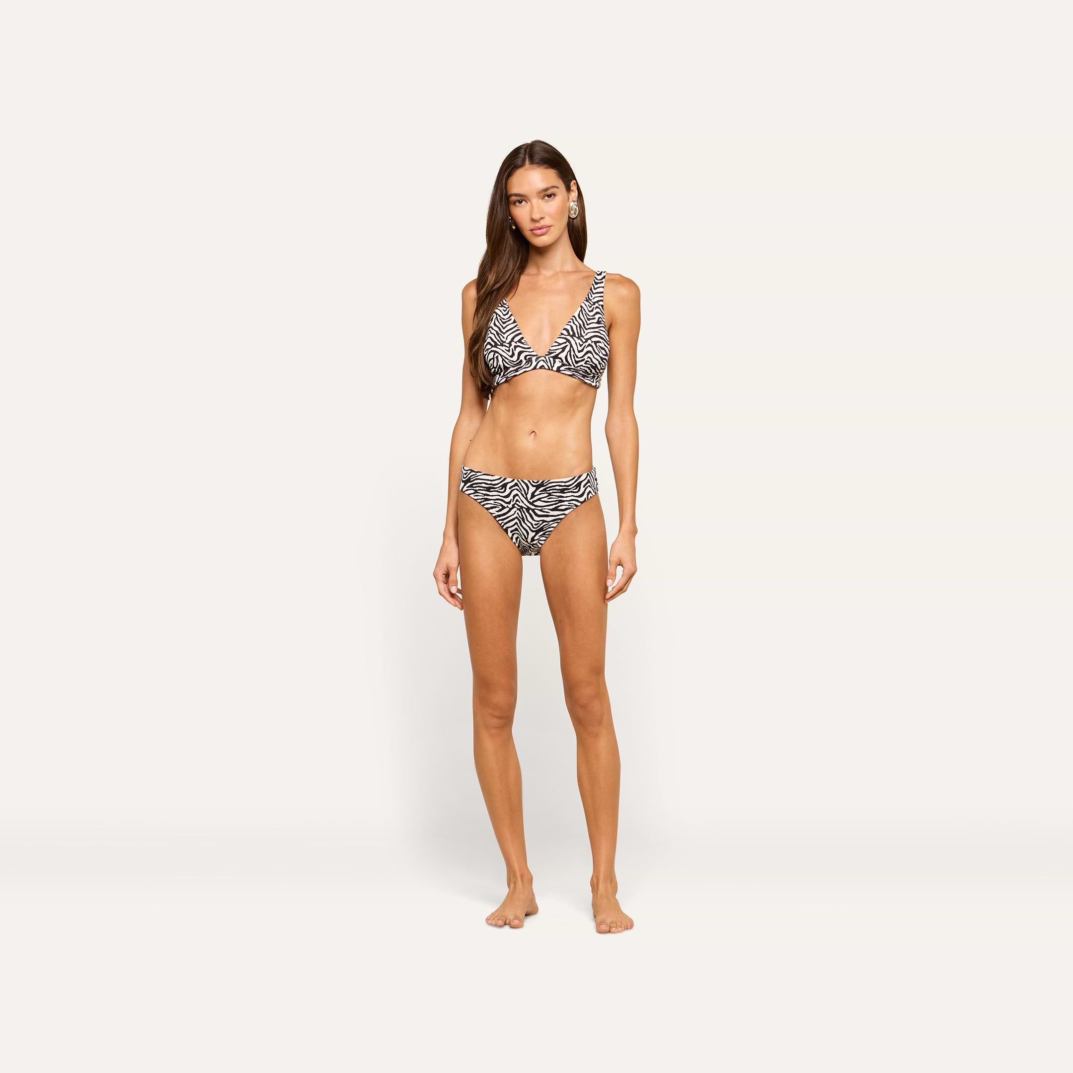 A woman with long brown hair stands barefoot against a plain light background, wearing a zebra print bikini and the Nicole Bikini Bottom, her arms relaxed at her sides and her expression neutral.