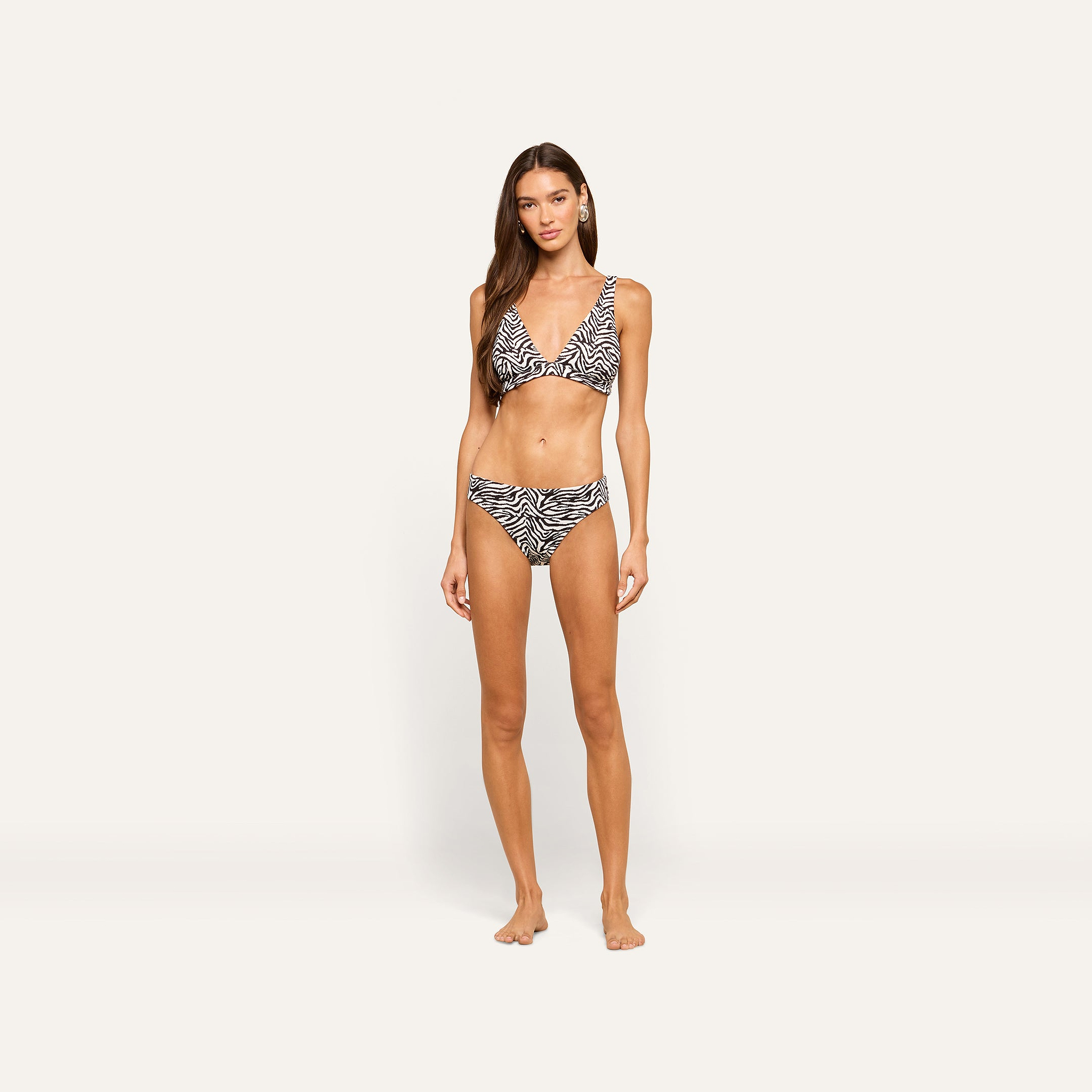 A woman with long brown hair stands barefoot against a plain light background, wearing a zebra print bikini and the Nicole Bikini Bottom, her arms relaxed at her sides and her expression neutral.