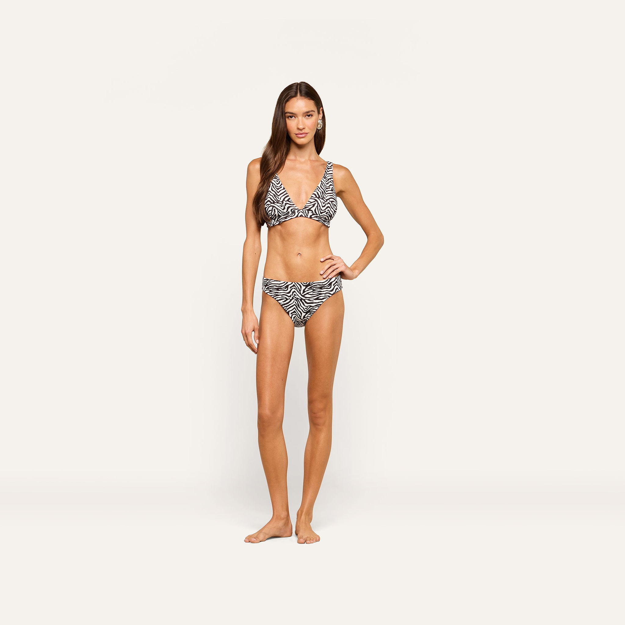 Barefoot woman poses against a plain light background, wearing a zebra print bikini with the Nicole Bikini Bottom, one hand on her hip and the other relaxed by her side.