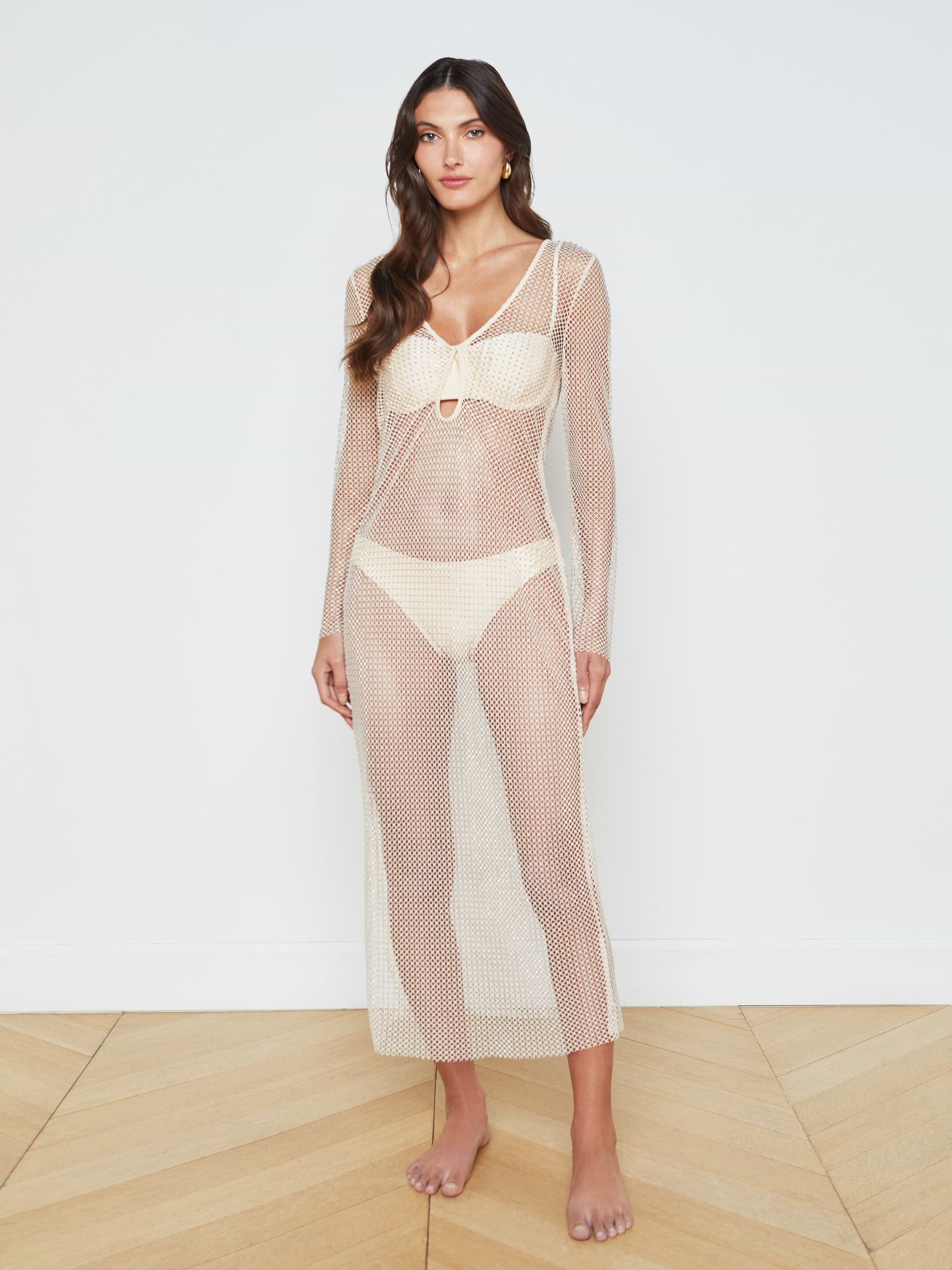 A woman stands barefoot against a plain white background, wearing the Sara Mesh Cover-Up Dress—a long, sheer mesh maxi dress with long sleeves—layered over a white bikini.