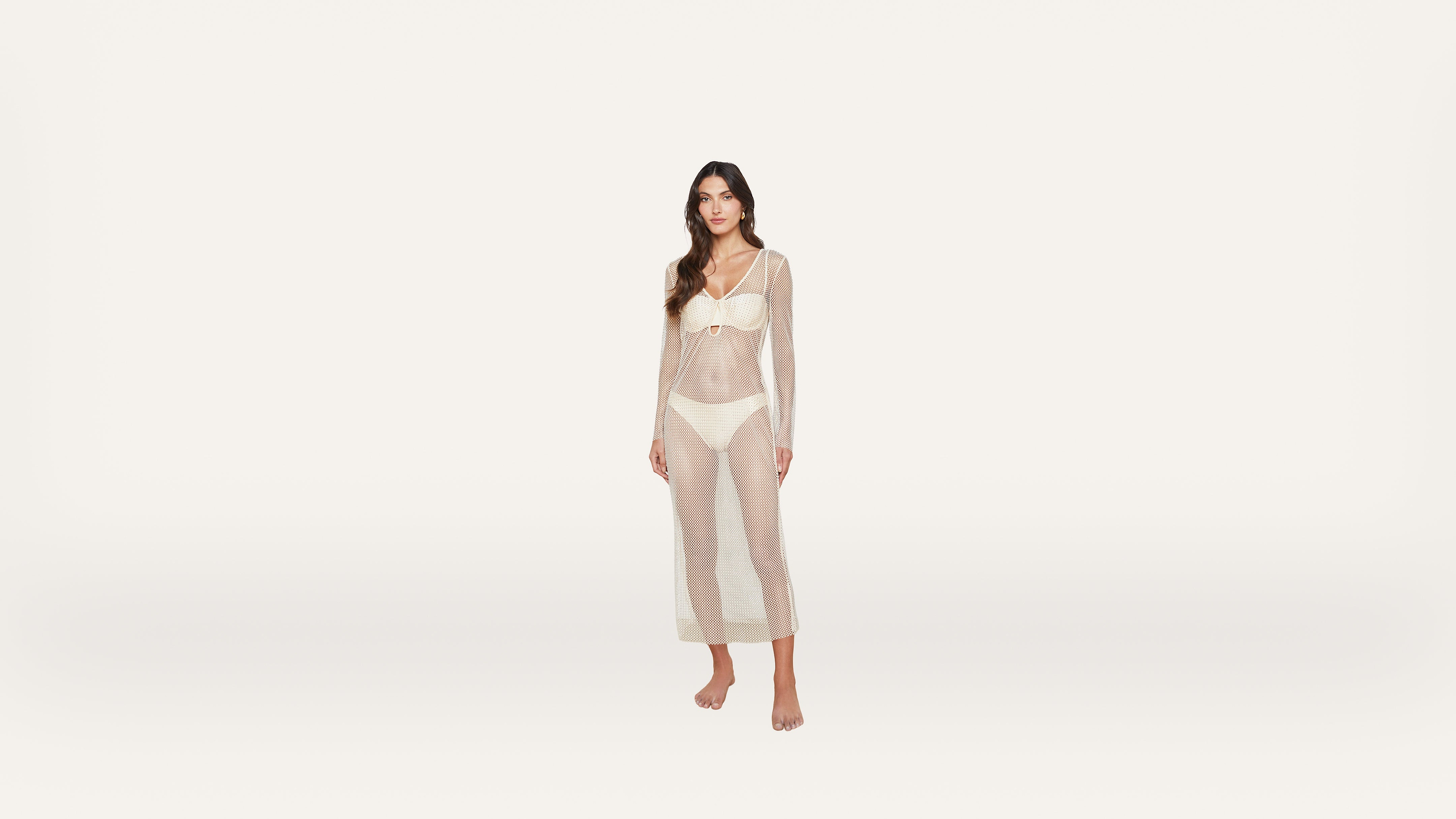 A woman stands barefoot in a cream bikini and a sheer, long-sleeved, ankle-length mesh cover-up dress against a plain white background.