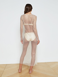 A woman stands barefoot, facing away, wearing the Sara Mesh Cover-Up Dress—a sheer mesh maxi dress with long sleeves and an ankle-length hem—layered over a light-colored bra and briefs, set against a plain white background.