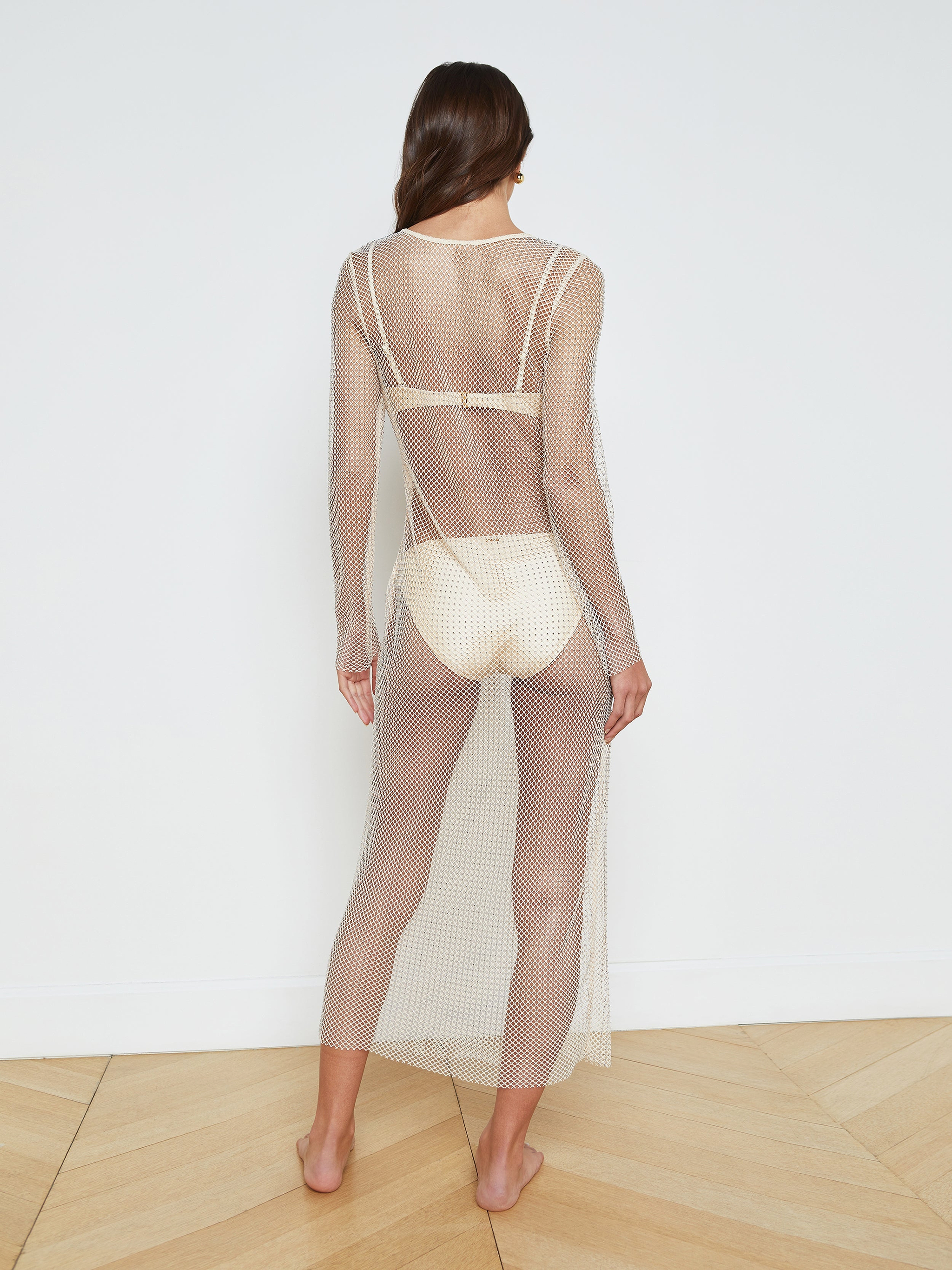 A woman stands barefoot, facing away, wearing the Sara Mesh Cover-Up Dress—a sheer mesh maxi dress with long sleeves and an ankle-length hem—layered over a light-colored bra and briefs, set against a plain white background.