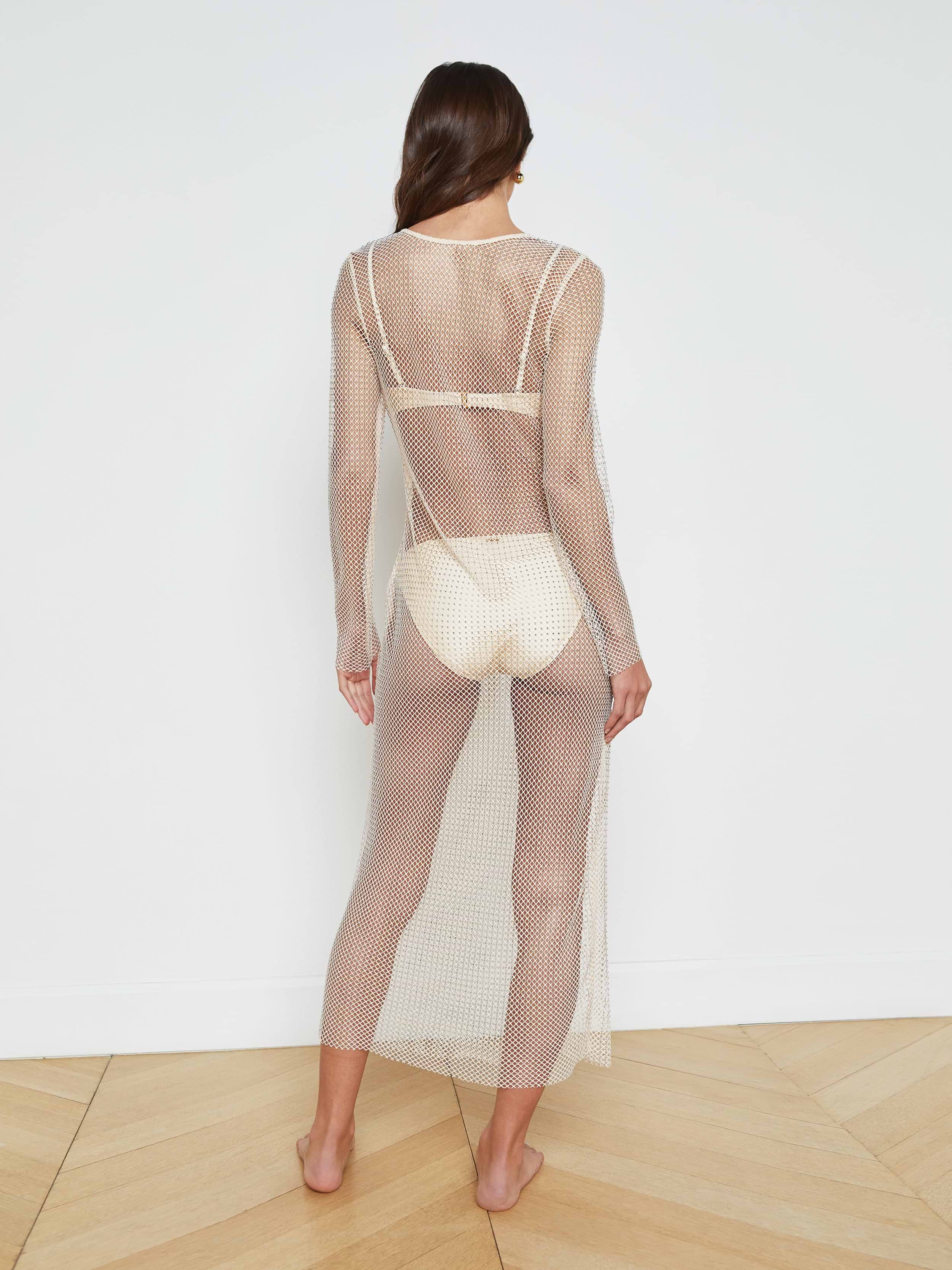 A woman stands barefoot, facing away, wearing the Sara Mesh Cover-Up Dress—a sheer mesh maxi dress with long sleeves and an ankle-length hem—layered over a light-colored bra and briefs, set against a plain white background.