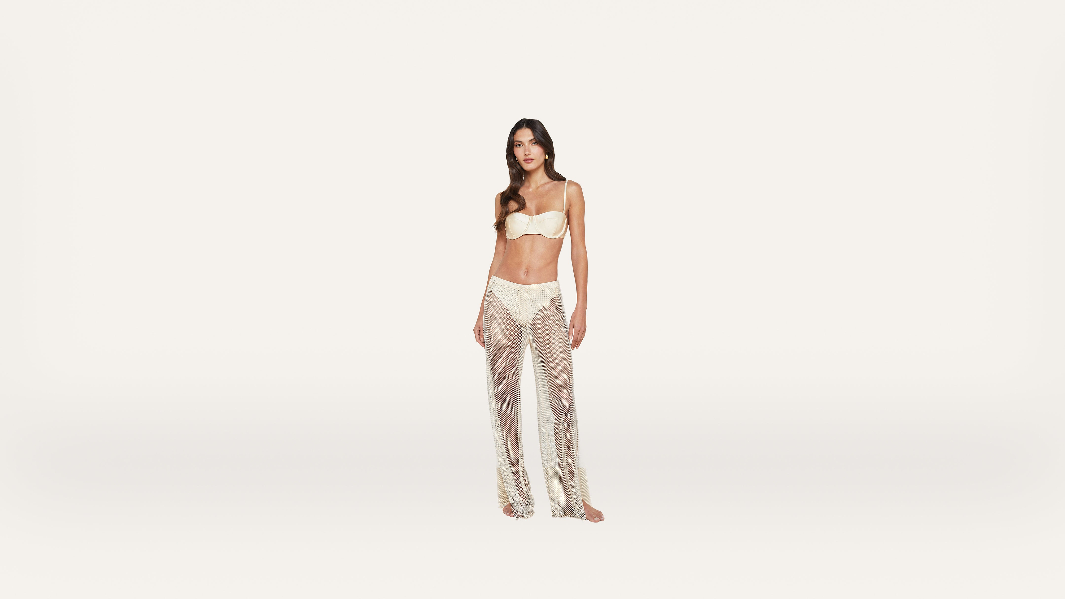 A woman stands on a white background wearing a cream-colored bra, matching underwear, and sheer, glittery pants.