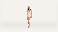 Woman in a cream-colored strapless swimsuit with a cutout, standing barefoot against a plain white background.
