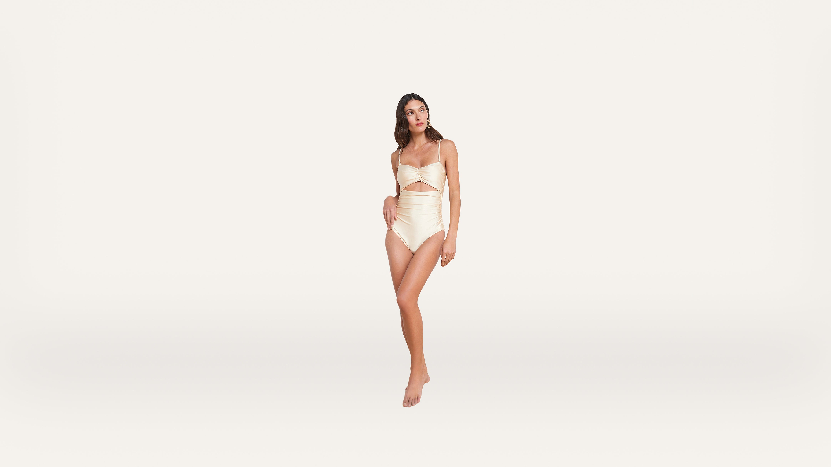 Woman in a cream-colored strapless swimsuit with a cutout, standing barefoot against a plain white background.
