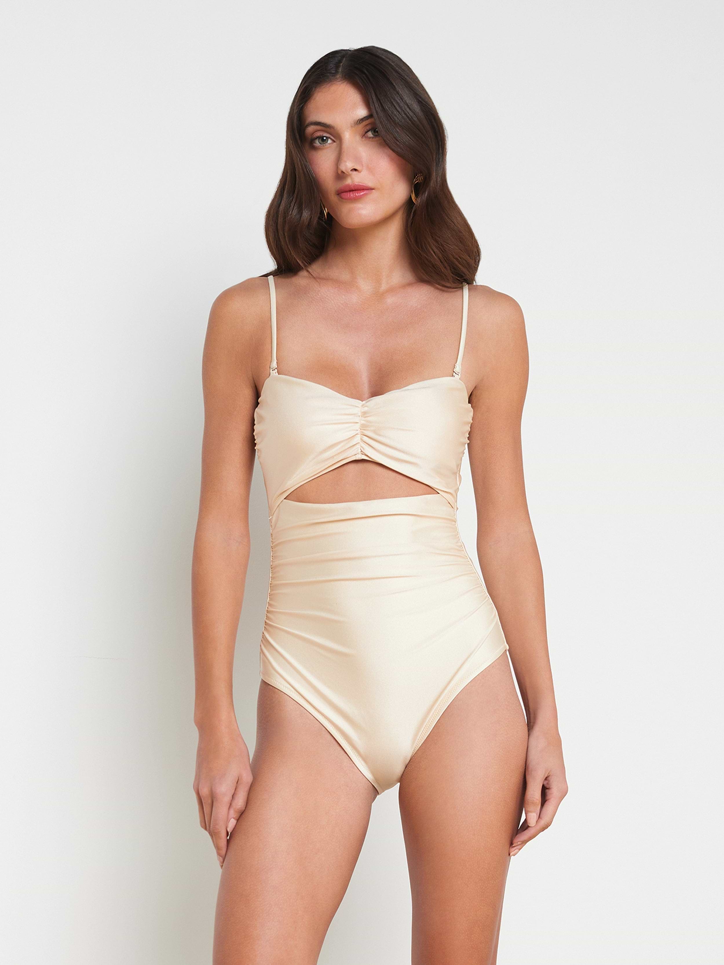 A woman wearing the Lily Cut-Out One-Piece Swimsuit—a light beige, high-waisted one-piece featuring a sweetheart neckline, ruched bodice, thin straps, and a cutout below the bust—stands against a plain off-white background.