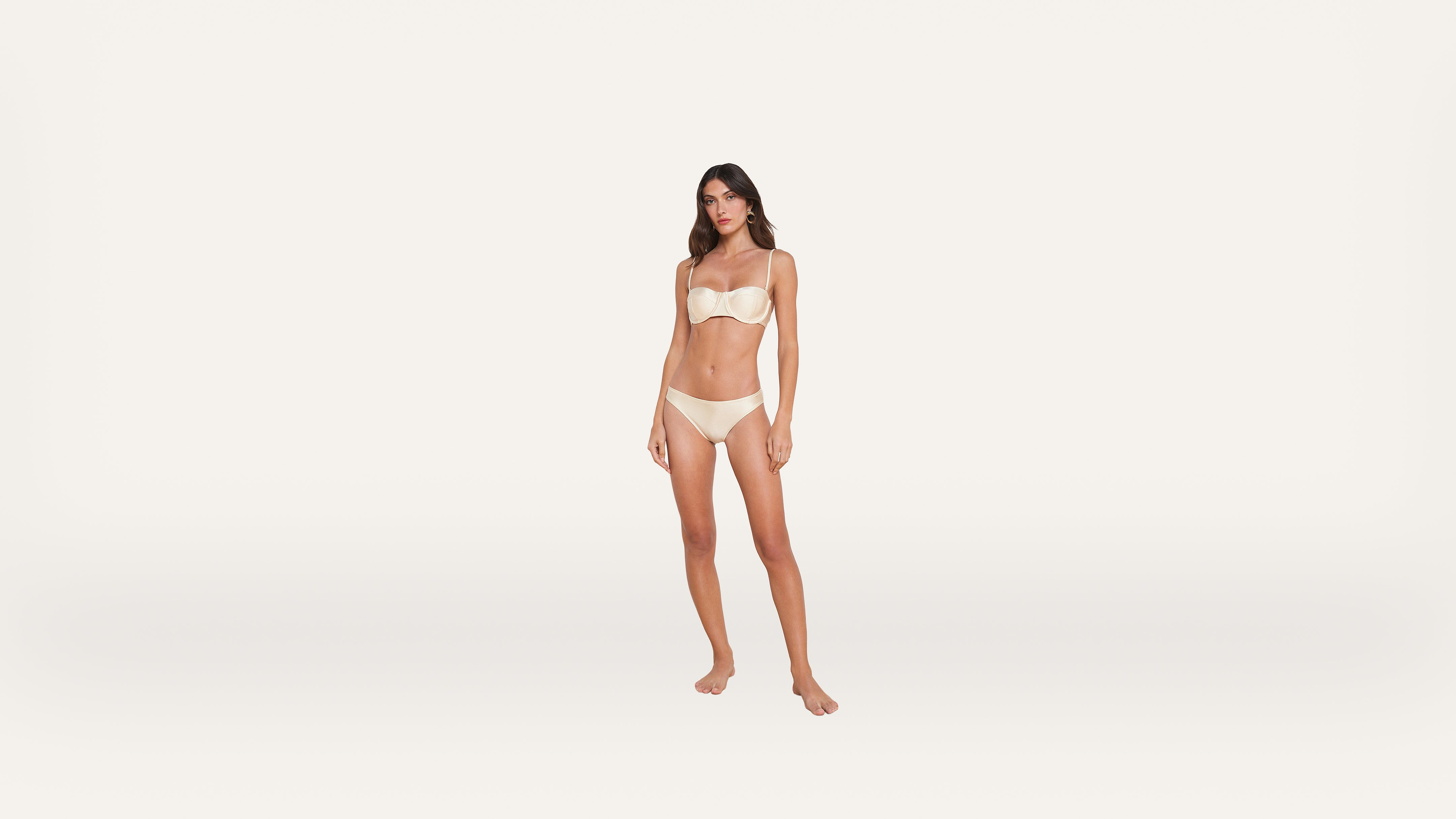 A woman stands barefoot against a plain white background, wearing a light-colored strapless bra and matching underwear.