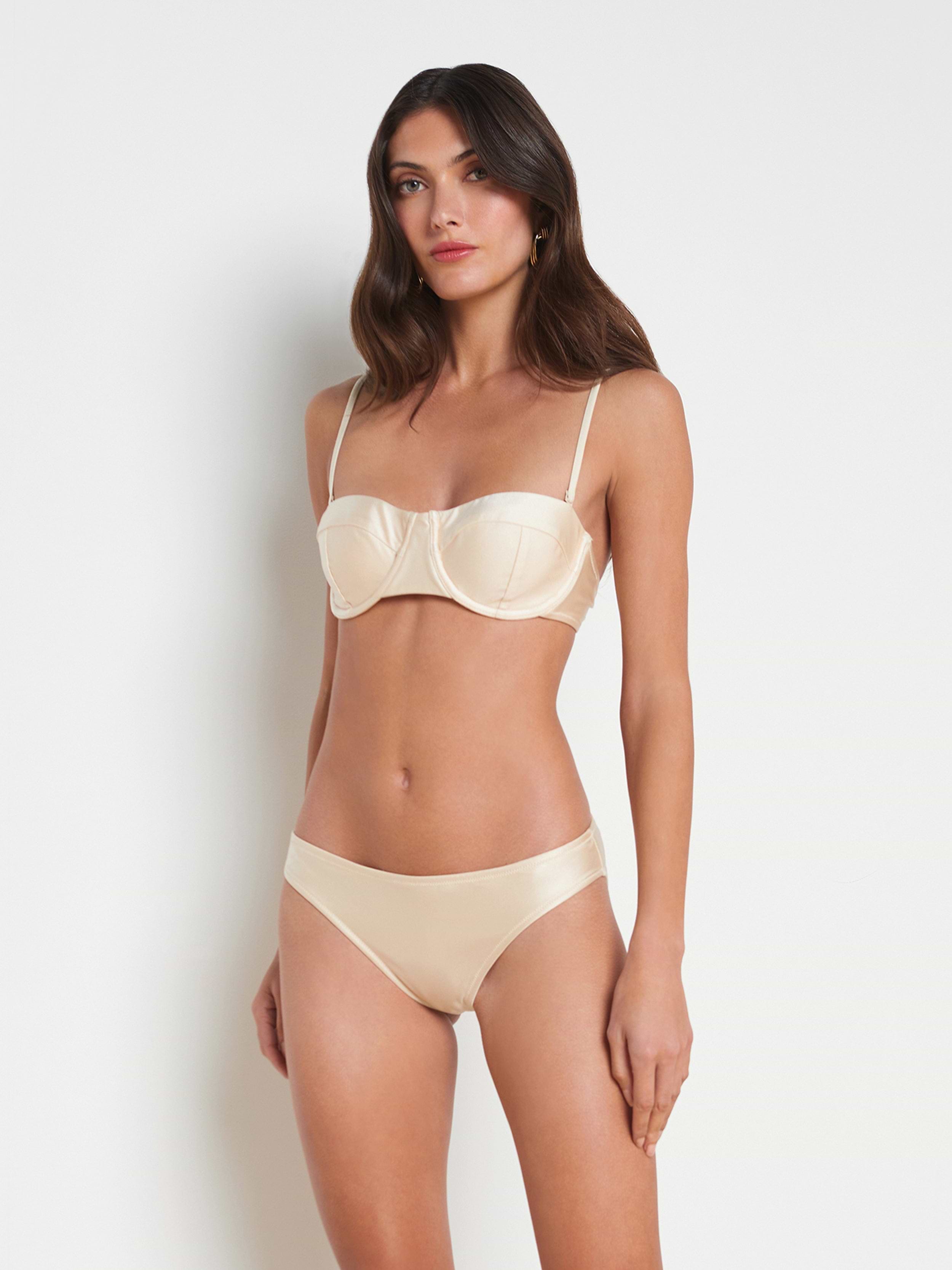 A woman stands against a plain background, wearing the Alexandria Bikini Top—a cream-colored underwire bikini with adjustable straps—paired with matching panties.
