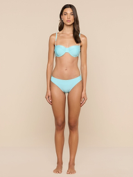 A woman stands facing forward against a plain background, wearing an ice blue Nicole Bikini Bottom made from stretch nylon and hoop earrings.