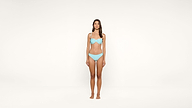 A woman stands barefoot wearing a light blue bikini in a minimal, white studio setting. She faces forward with a neutral expression and her arms at her sides.