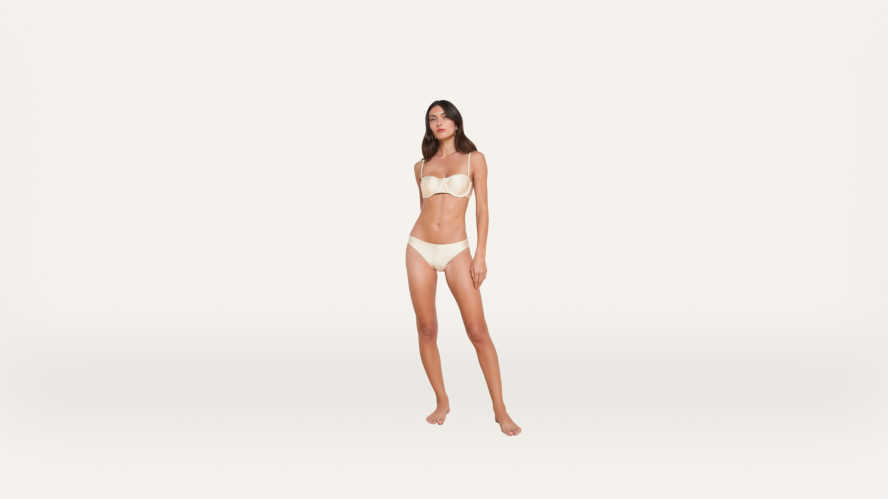 A woman stands barefoot against a plain white background, wearing a matching strapless bra and underwear set in a light beige color. She faces forward with arms relaxed at her sides.