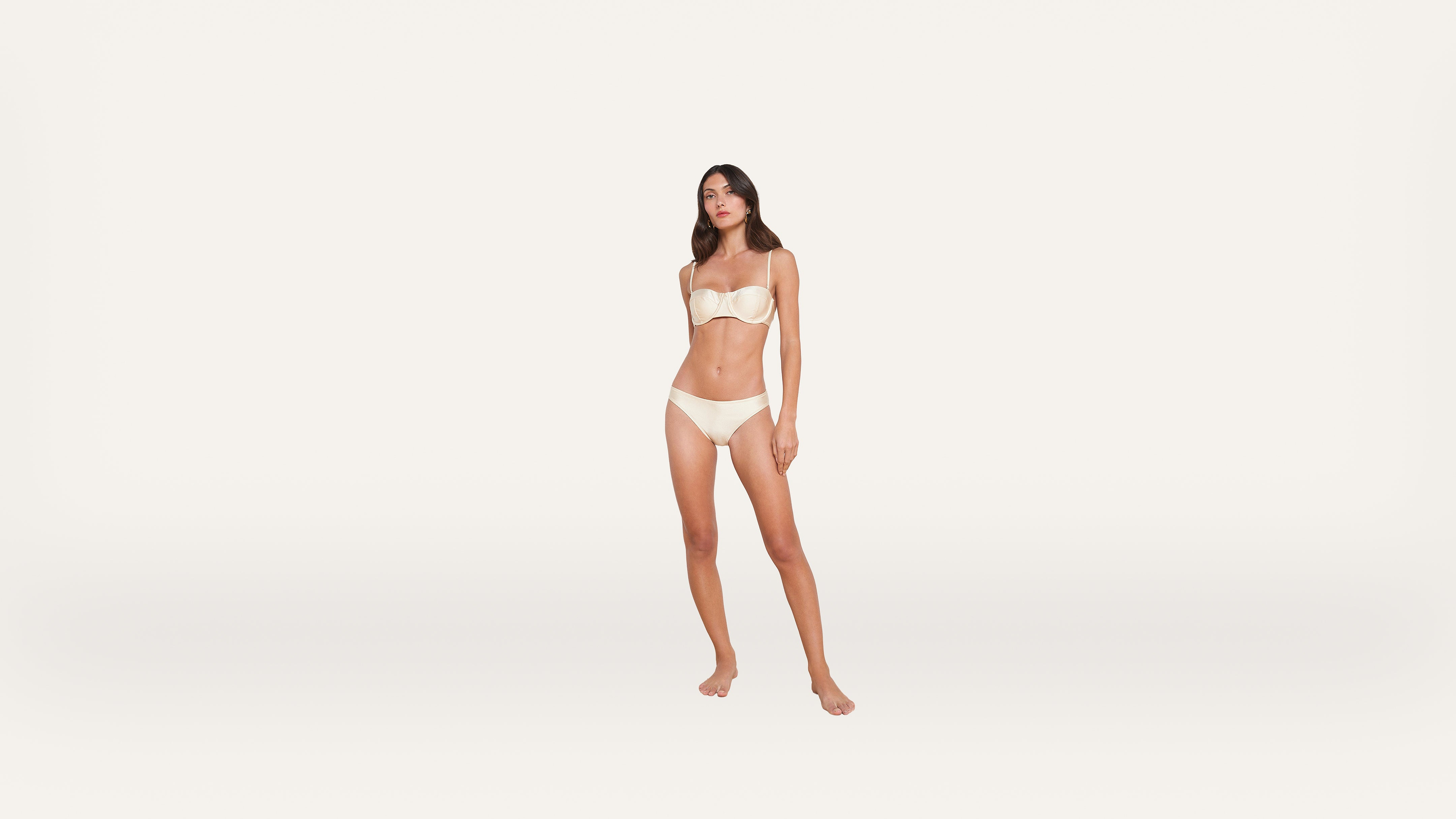 A woman stands barefoot against a plain white background, wearing a matching strapless bra and underwear set in a light beige color. She faces forward with arms relaxed at her sides.