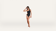 Woman wearing sunglasses and a black one-shoulder swimsuit poses against a plain white background.