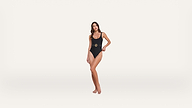 A woman stands barefoot wearing a black one-piece swimsuit with a belt detail, against a plain white background.