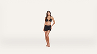 Woman standing barefoot against a plain white background, wearing a black bikini top and a black mini skirt, looking slightly to the side with one hand on her hip.