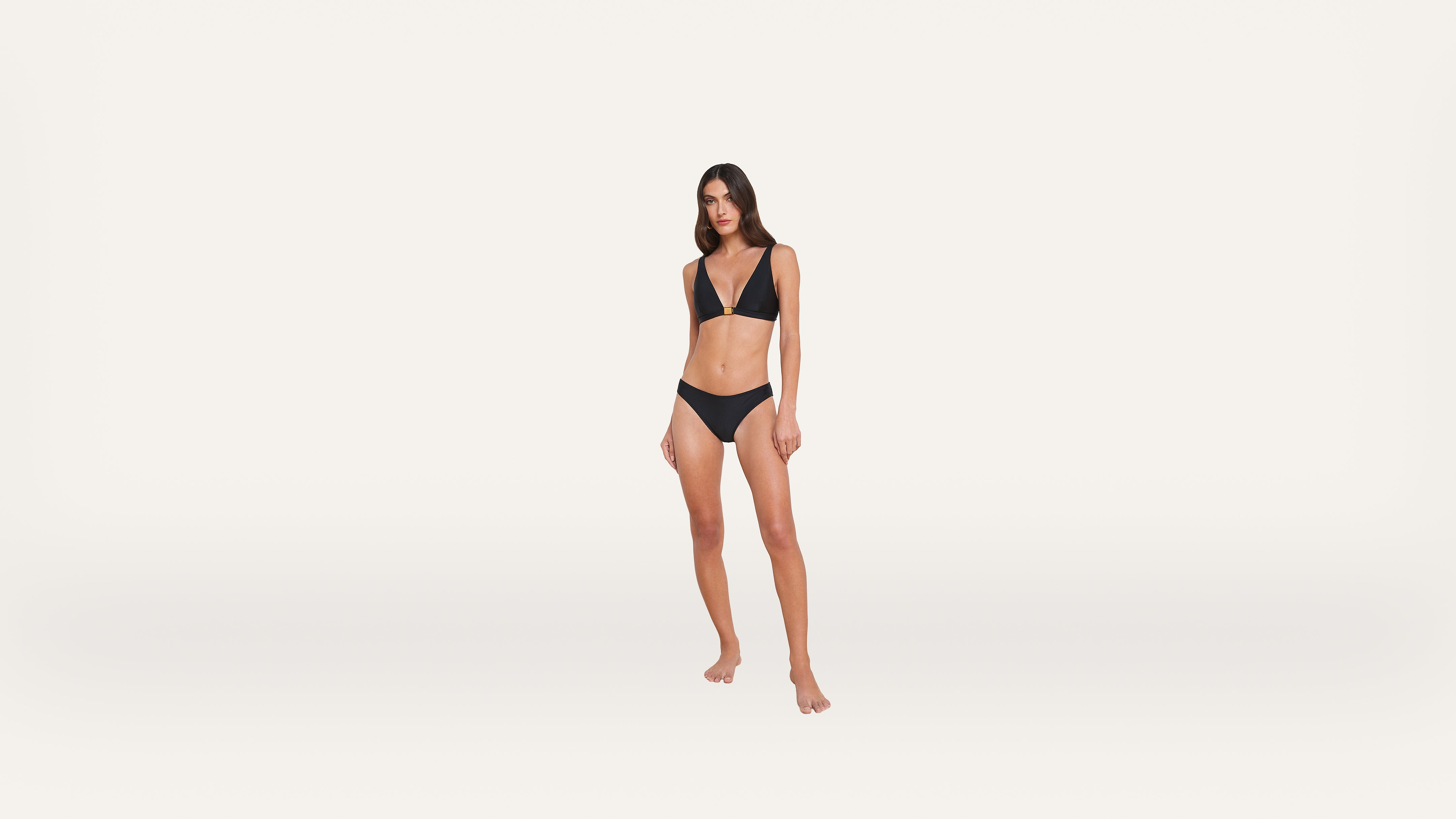 A woman stands barefoot against a plain white background, wearing a black bikini.