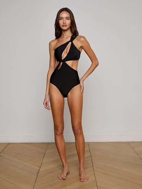 A woman stands barefoot against a plain white background, wearing the Camilla Monokini—an elegant black one-shoulder swimsuit featuring stylish cutout details.
