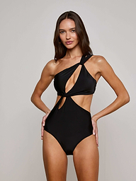 A woman models the Camilla Monokini—a black, one-shoulder swimsuit with a cutout design—posing with her hands on her hips and smiling against a plain light background.
