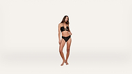 A woman stands barefoot against a plain white background, wearing a black bikini. She has long hair and a relaxed, confident pose, with one arm bent and the other hanging by her side.