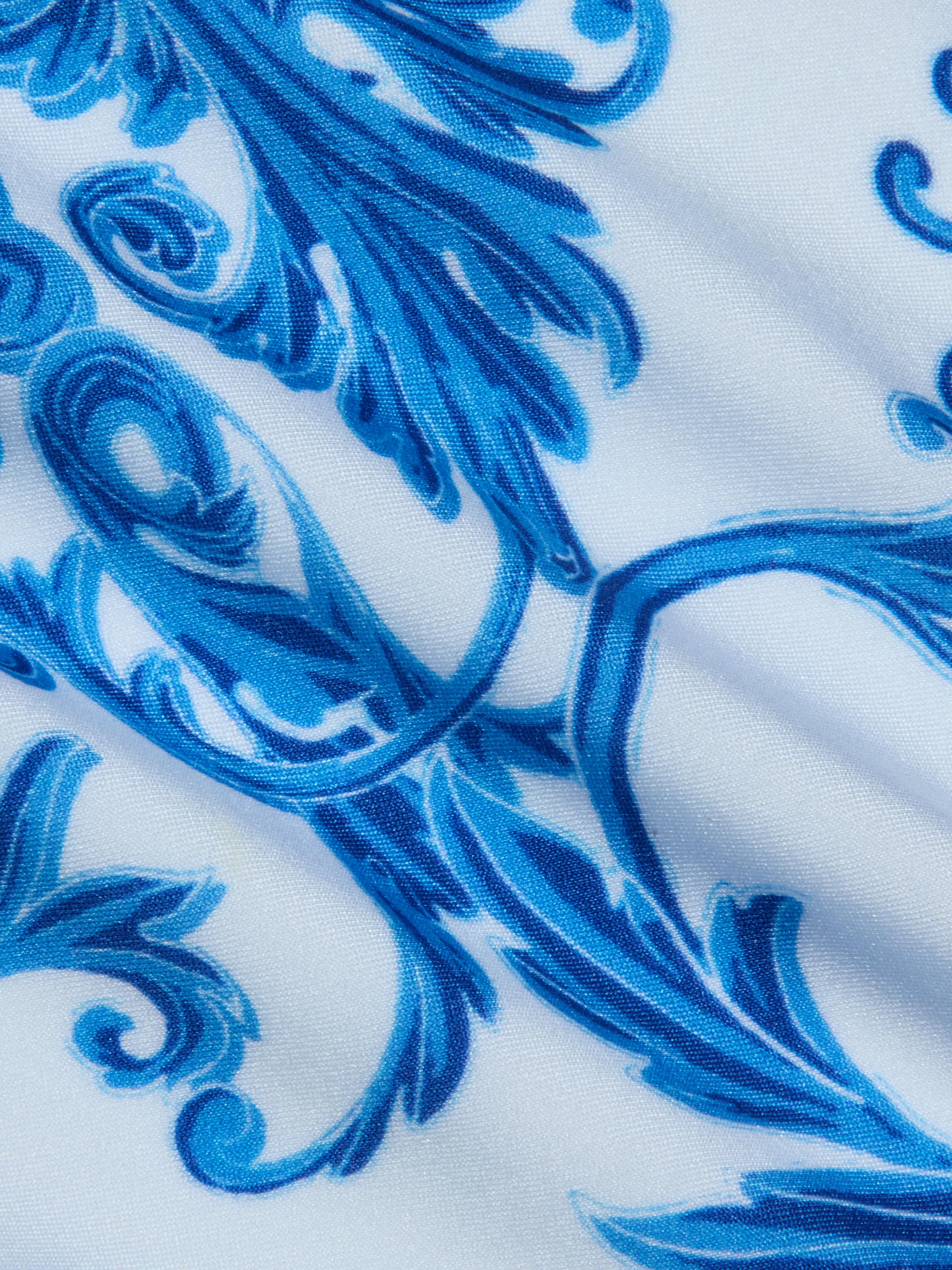 Close-up of the Remi One-Piece Swimsuit, showcasing its white fabric adorned with a blue ornate floral pattern. The design features swirling and leaf-like baroque-inspired motifs, while subtle wrinkles in the fabric enhance the textured look of this decorative swimwear.