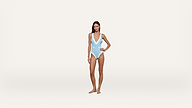 Woman standing against a white background, wearing a light blue one-piece swimsuit with a deep V neckline and white trim, posing with one hand on her hip and the other relaxed by her side.