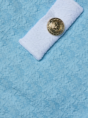 A white towel with a gold button sits atop a larger light blue textured towel, echoing the powder blue color of the Lisa Plunge One-Piece Swimsuit. The background is fully covered by the blue towel.