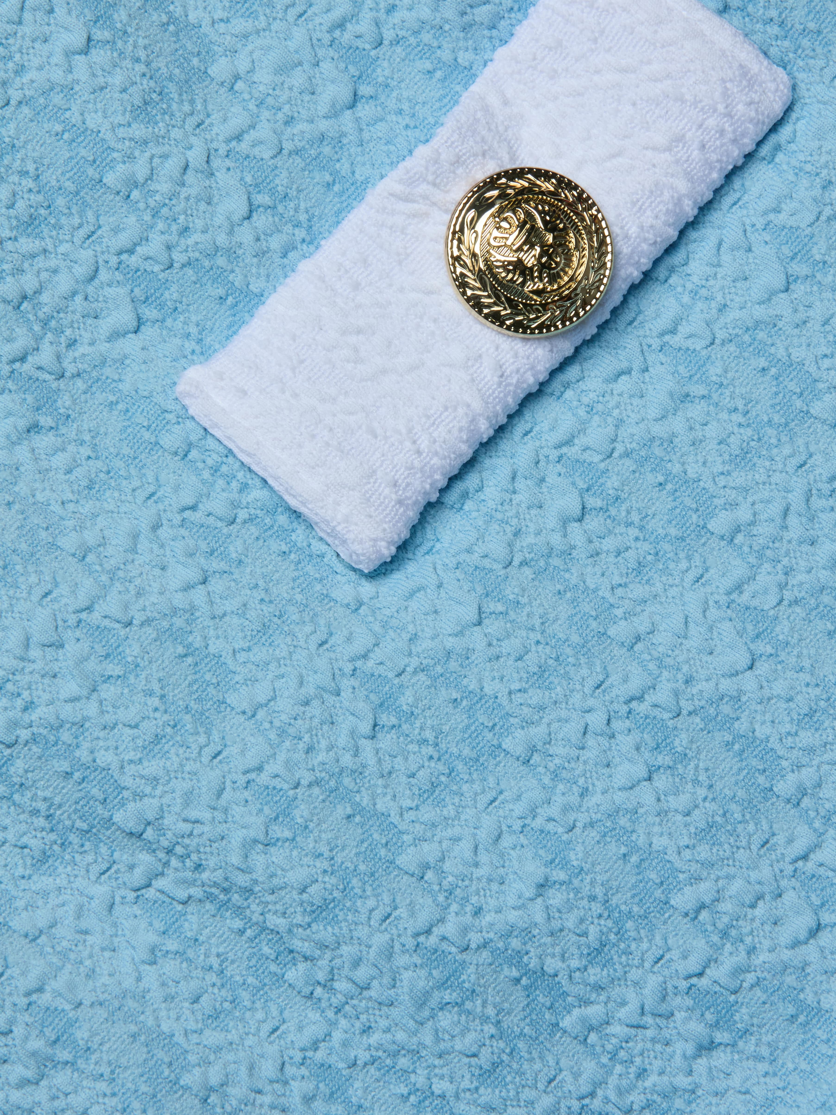 A rolled white towel with a gold decorative emblem rests on a light blue textured surface, evoking the soft bouclé swim fabric of the Lisa Plunge One-Piece Swimsuit in powder blue.