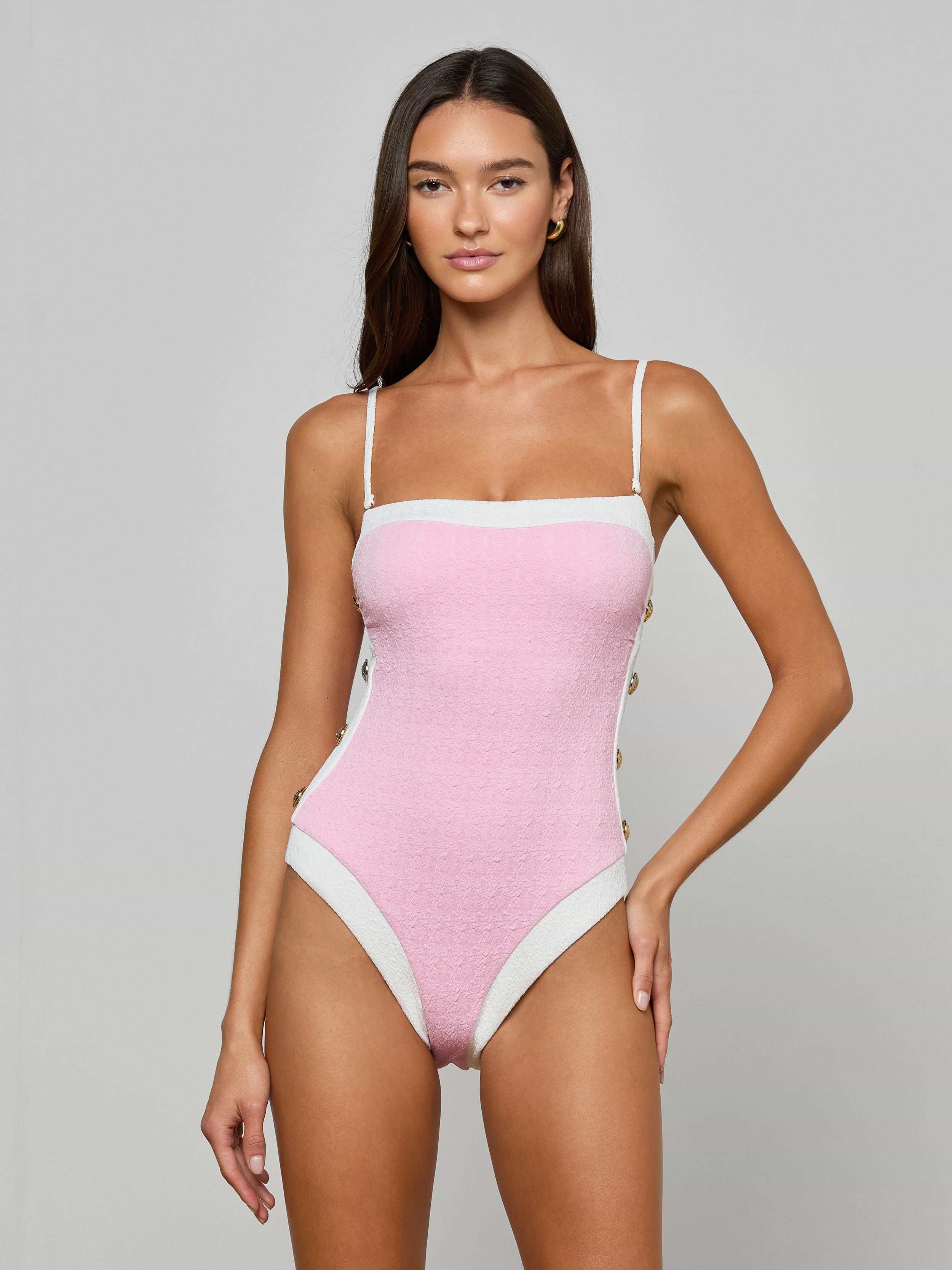 A woman stands against a plain light background, wearing the Tory Bandeau One-Piece Swimsuit—a pink, bouclé fabric one-piece with white trim and thin straps.
