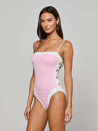 A woman models the Tory Bandeau One-Piece Swimsuit, crafted from pink bouclé fabric with white straps and panels, and accented by gold buttons on the side. She stands against a plain light background, gazing confidently at the camera.