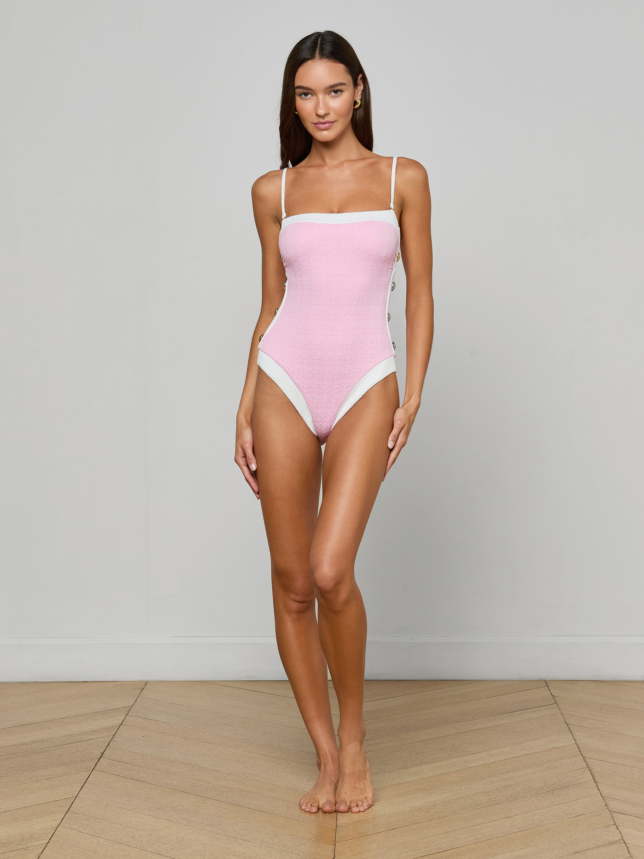 A woman stands barefoot against a plain white background, wearing the Tory Bandeau One-Piece Swimsuit in light pink with thin straps and white trim details, looking directly at the camera with a neutral expression.